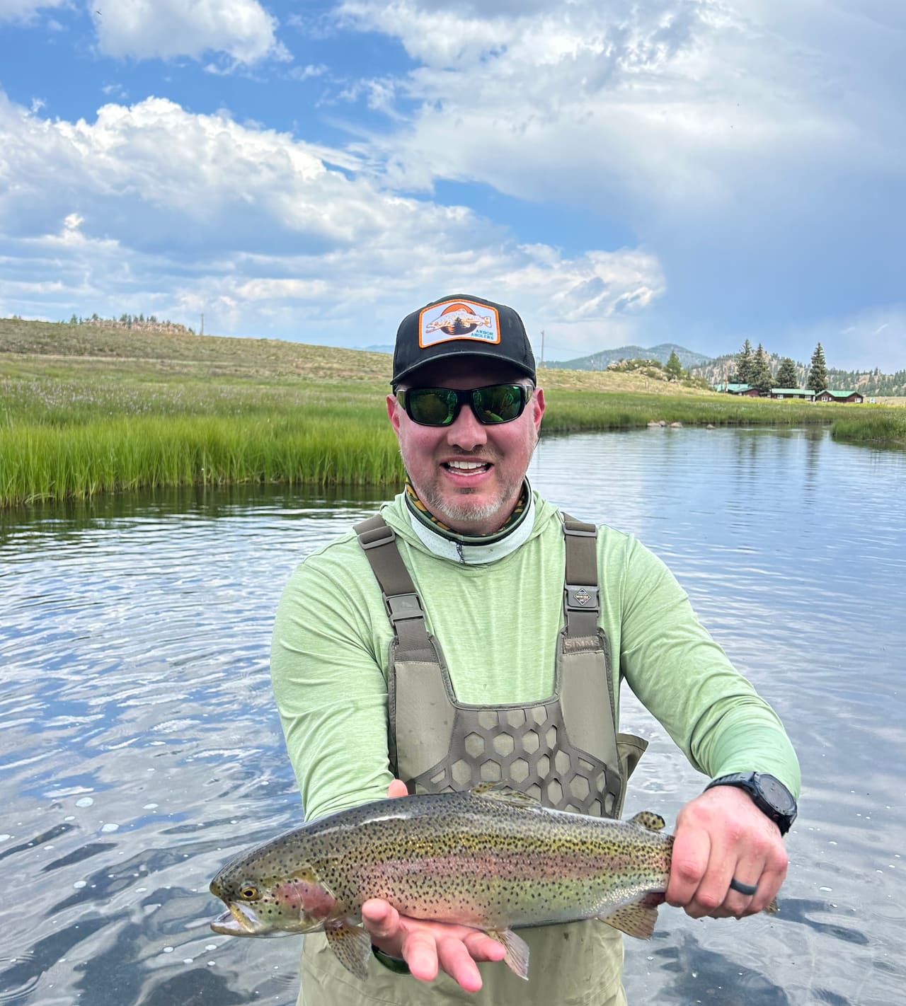 An angler in waders holding a rainbow trout on a high meadow stream under big clouds.